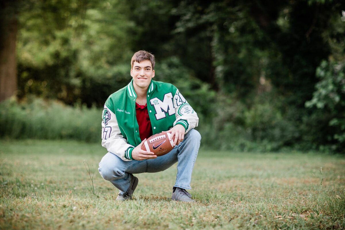 High school football player squatting down in green grass wearing his letter jacket and holding his ball.