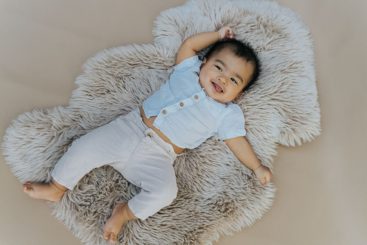 Smiling baby boy dressed in a light blue shirt and light pants, lying on a soft beige fur rug with one arm playfully stretched.