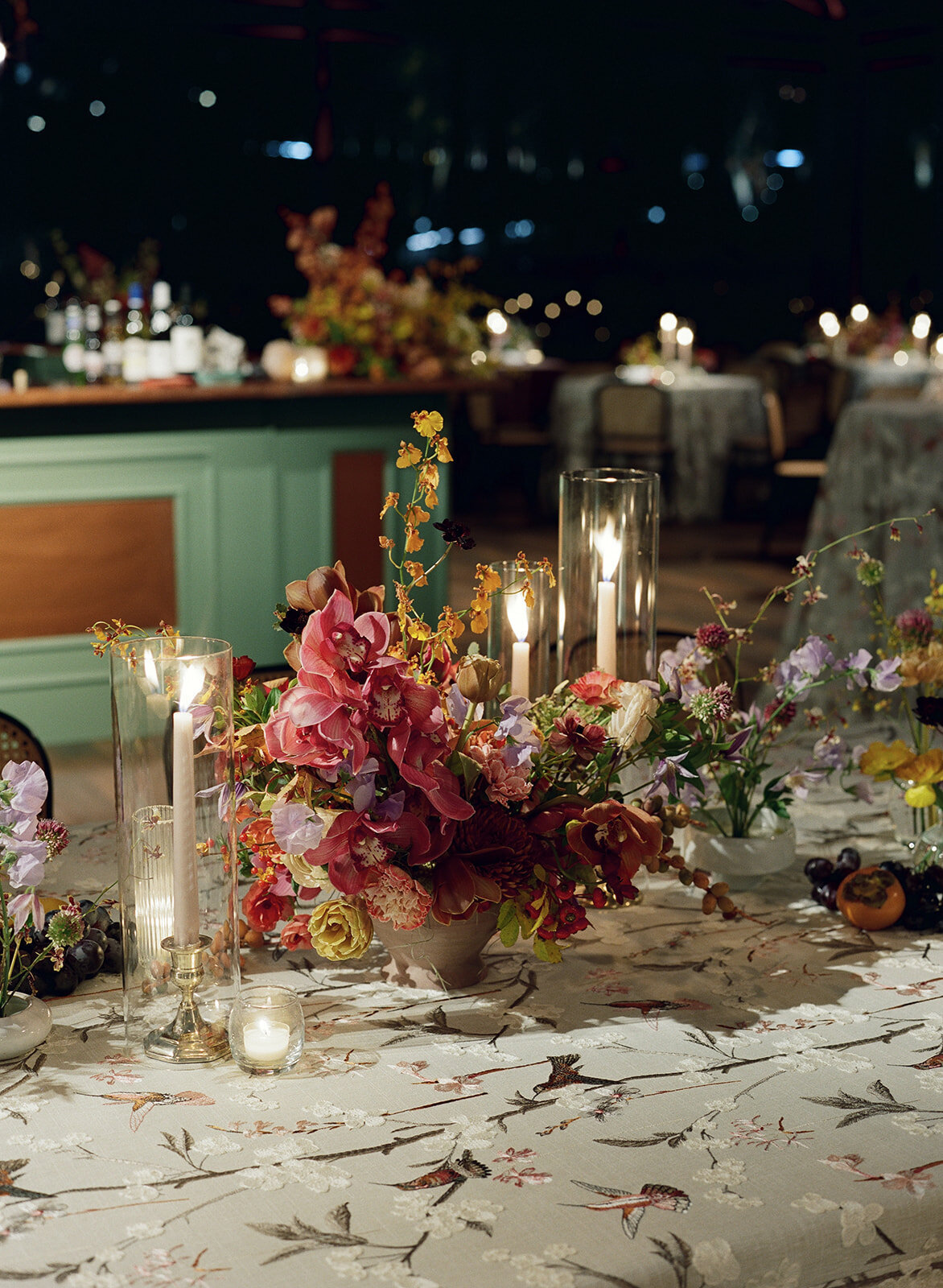 Clustered and styled floral arrangements, fruits, and art pieces for fall wedding reception. Low centerpieces mixed with ikebana arrangements and bud vases decorate the reception tables at this fall wedding. Floral centerpieces featuring orchids, ranunculus, sweet peas, and bittersweet. Styled wedding tables with fruits, curated candles, and floral arrangements. Tasteful fall color palette of copper, ruby, gold, burgundy, rust, and lavender. Design by Rosemary & Finch Floral Design in Nashville, TN.