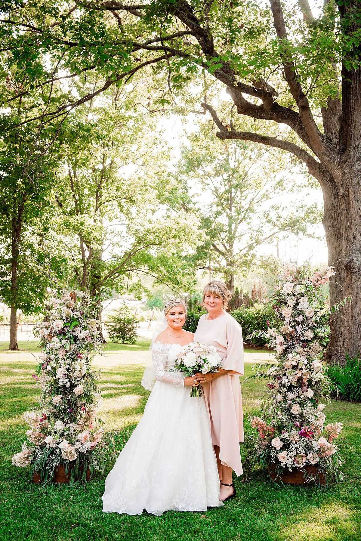A bride wearing an off the shoulder long sleeved lace wedding dress stands next to her mother wearing a blush long sleeved dress holding a large bouquet of white peonies and blush roses. They stand in front of the lush floral arbor bursting with pampas grass, eucalyptus, blush roses, ivory roses, white hydrangea and greenery at Arrington Vineyards wedding