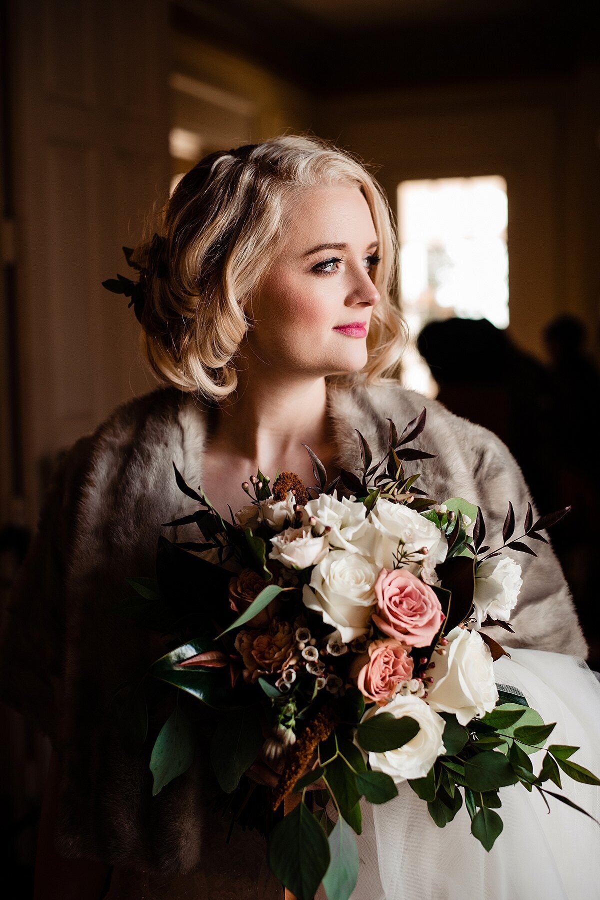 A bride with short blonde hair in finger waves looks out the window of Rippa Villa on her wedding day. The bride is wearing a white tulle gown with a light brown fur wrap while holding a bouquet of white roses, blush roses and greenery wrapped in a velvet rust colored ribbon.