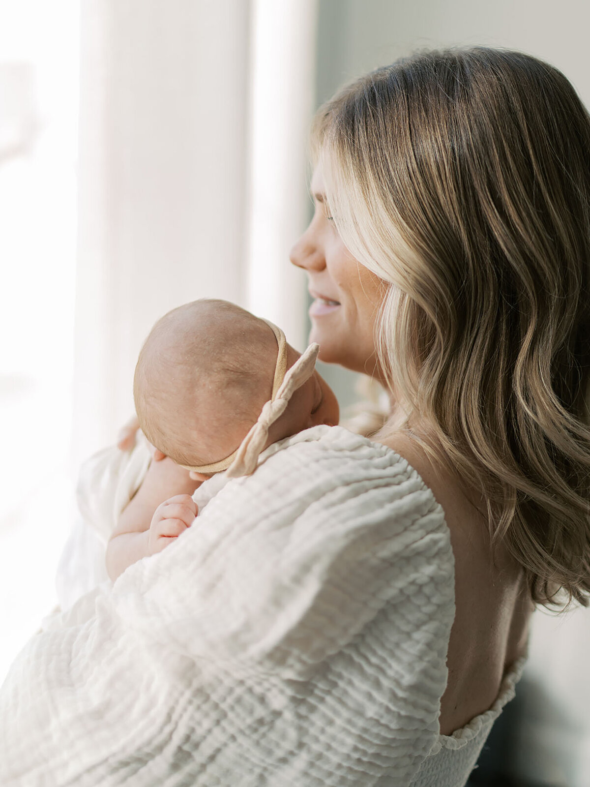 A new mother stands by the window, gently holding her newborn in her arms while gazing outside. The soft natural light highlights the love and serenity of this quiet, intimate moment.