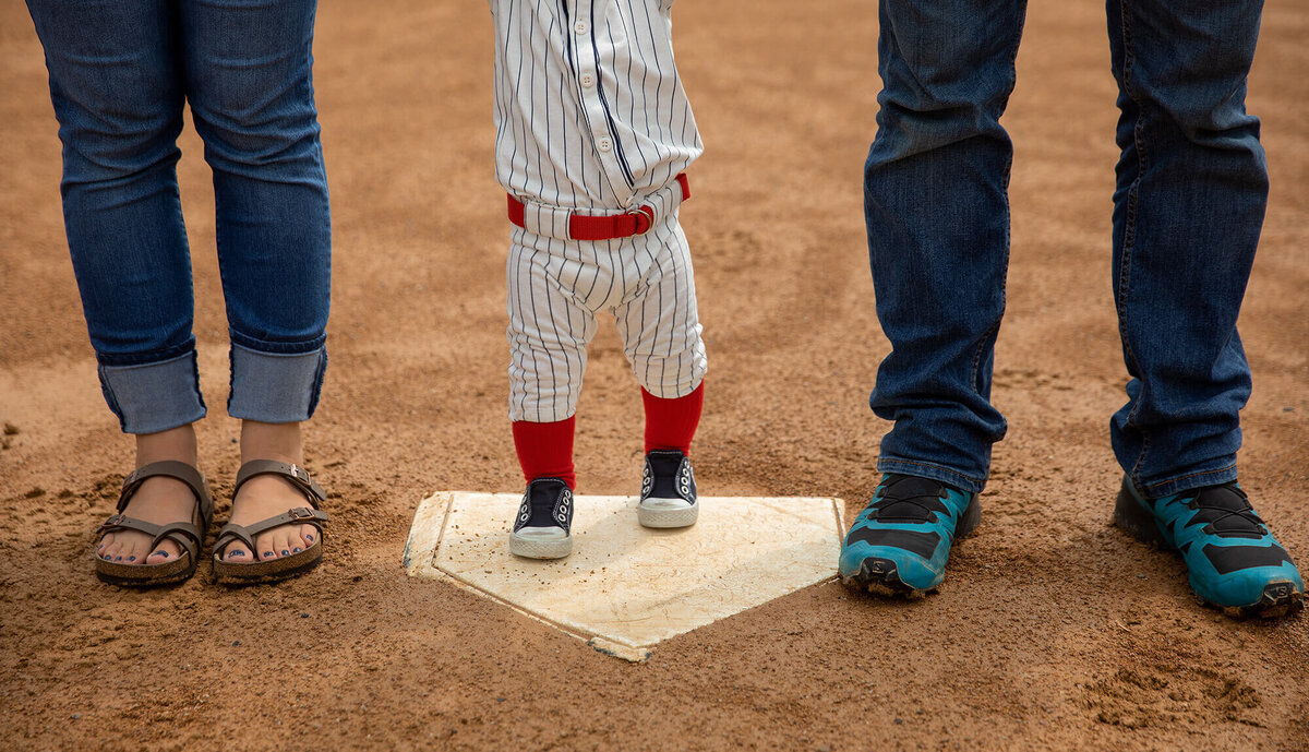 Legs of Dad, Mom and 1 year old boy with little boy standing on home plate.