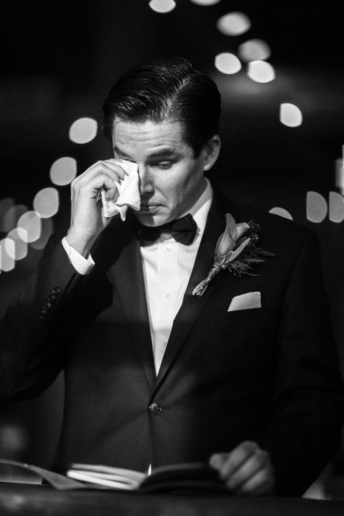 A groom emotionally wipes a tear from his eyes during a speech, set against a backdrop of soft, glowing lights.