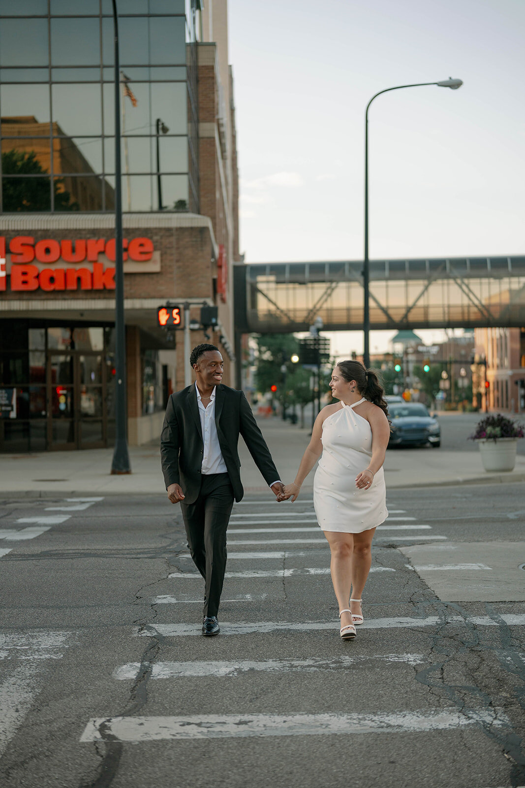 Downtown Kalamazoo crosswalk engagement photo of couple laughing and walking together.