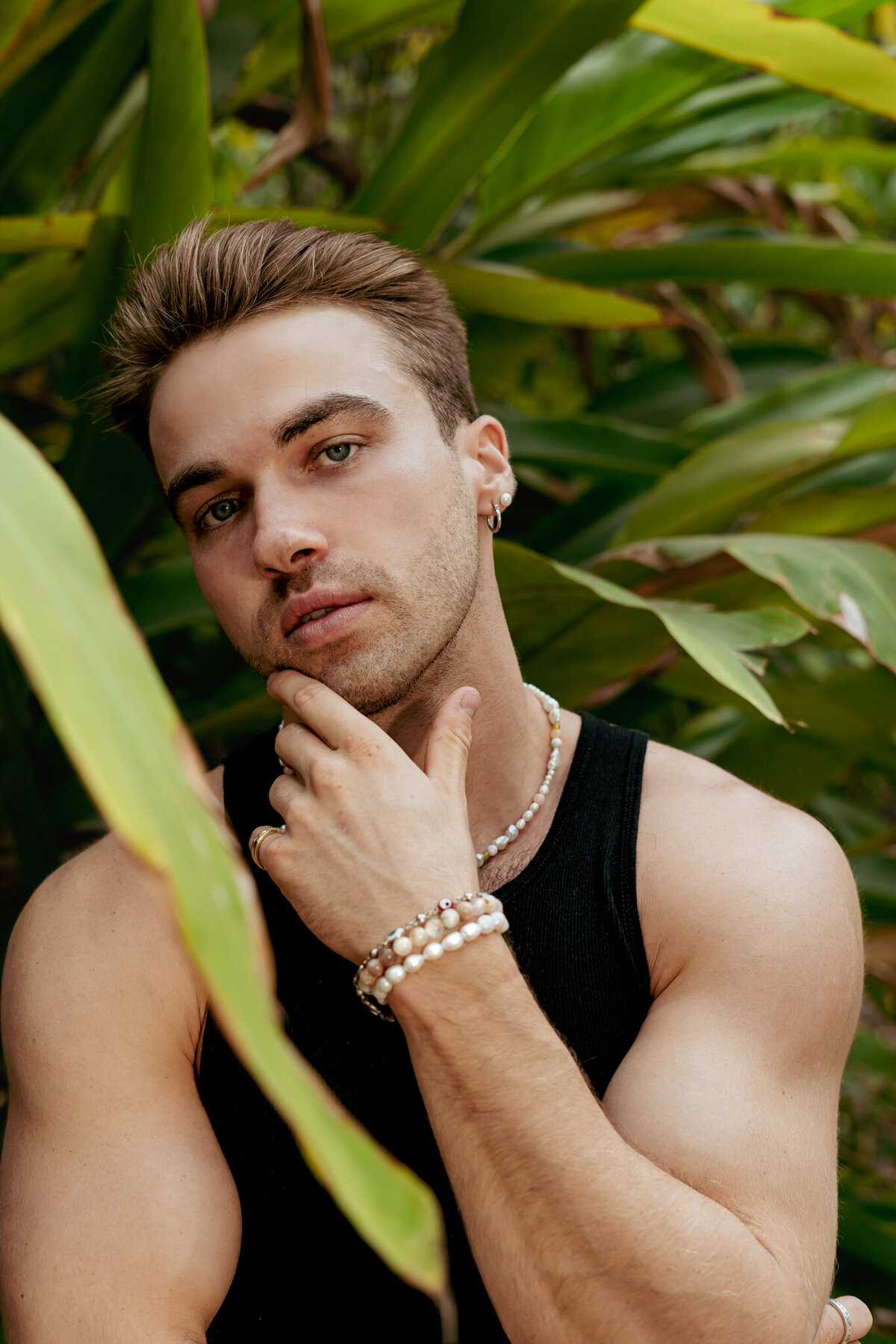 male model poses up close in portrait showing pearl bracelets while standing in the middle of green trees and forest