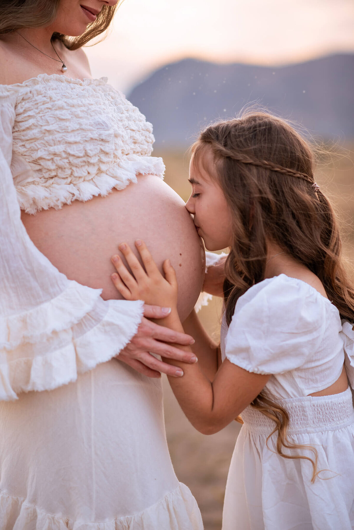 A little girl with long brown hair crouches down to embrace and kiss her pregnant mom's exposed belly.