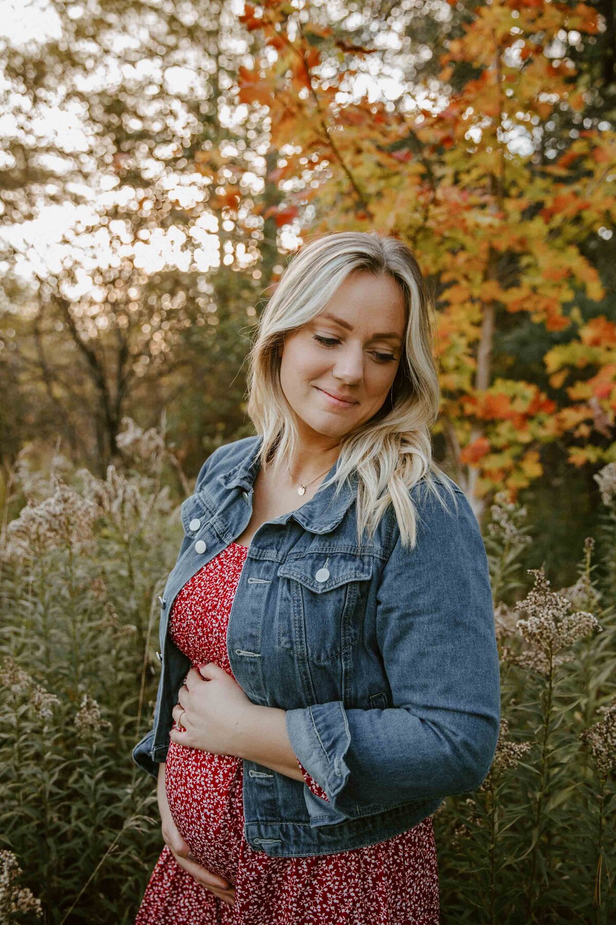 Expectant mom in outdoor maternity photo session in London, Ontario. Mom is waring a red patterned dress with denim jacket holding her baby bump and looking down over her shoulder.