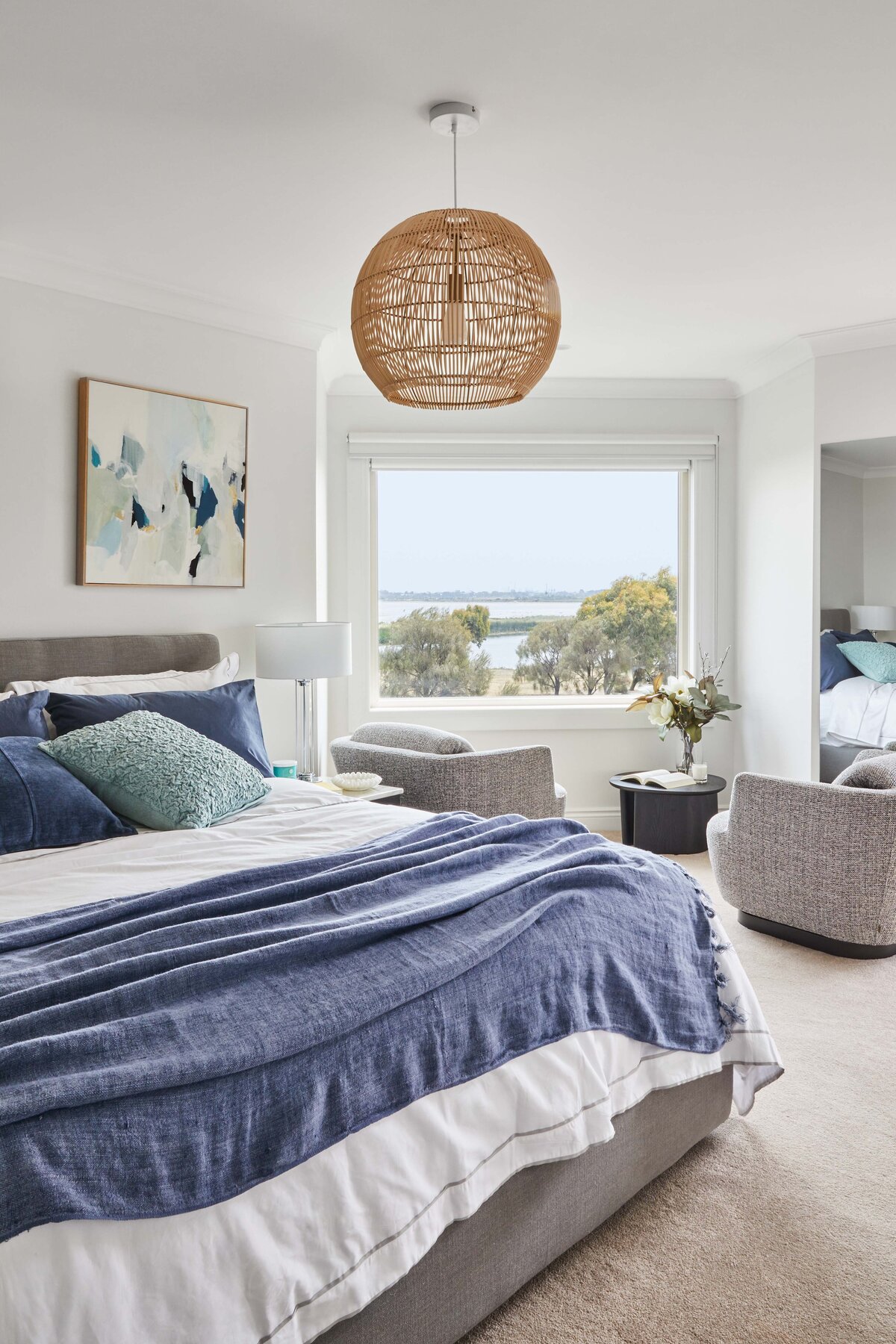 A spacious bedroom in a Williamstown home with a stunning water view from a large window. The room features a gray bed with blue bedding, a wicker pendant light, and a sitting area with two textured gray armchairs.