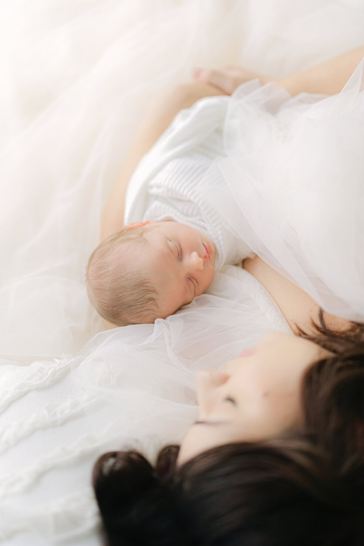 A mother lays peacefully in a white tulle dress with her sleeping newborn baby swaddled in white wrapped in her arms, Indianapolis Newborn Photographer