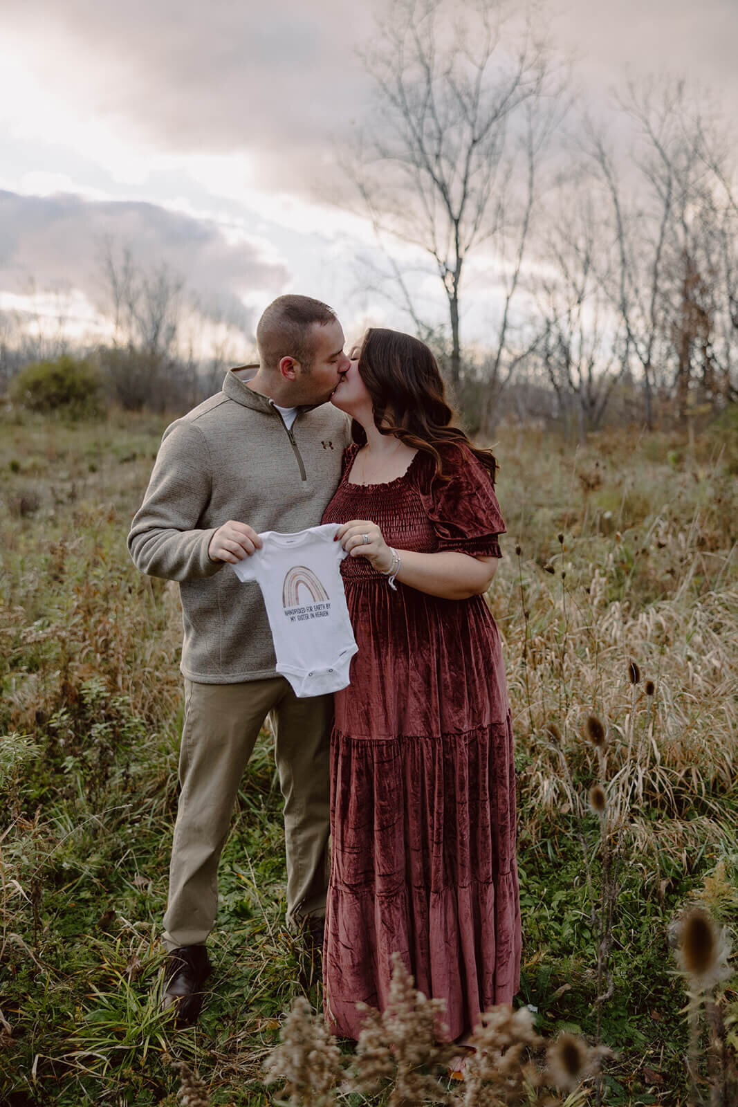 Couple kissing holding a baby onesie at  Finger Lakes Maternity session