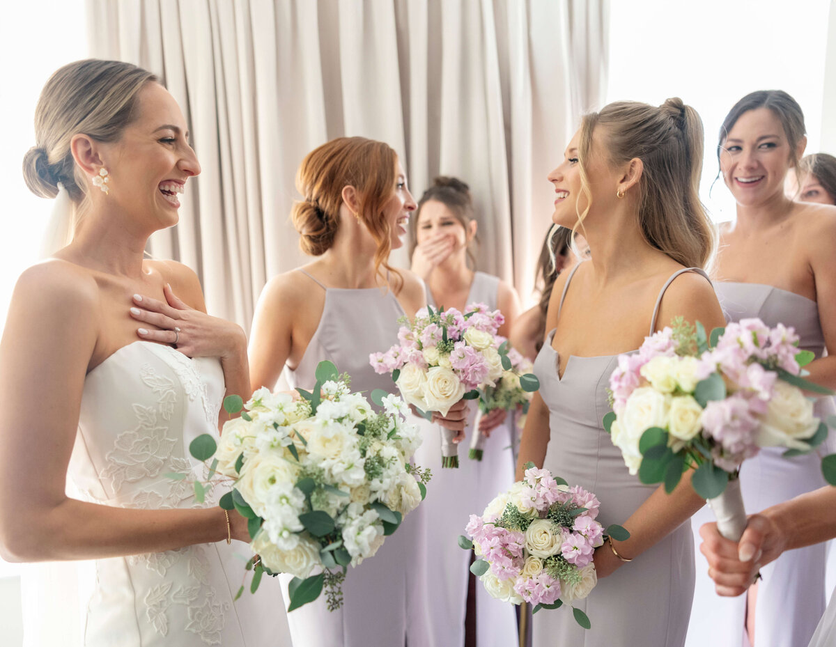 A joyful bride in a white gown holds a bouquet while surrounded by bridesmaids in lavender dresses, all smiling and holding floral bouquets. They stand near soft curtains, sharing a happy moment before the wedding.