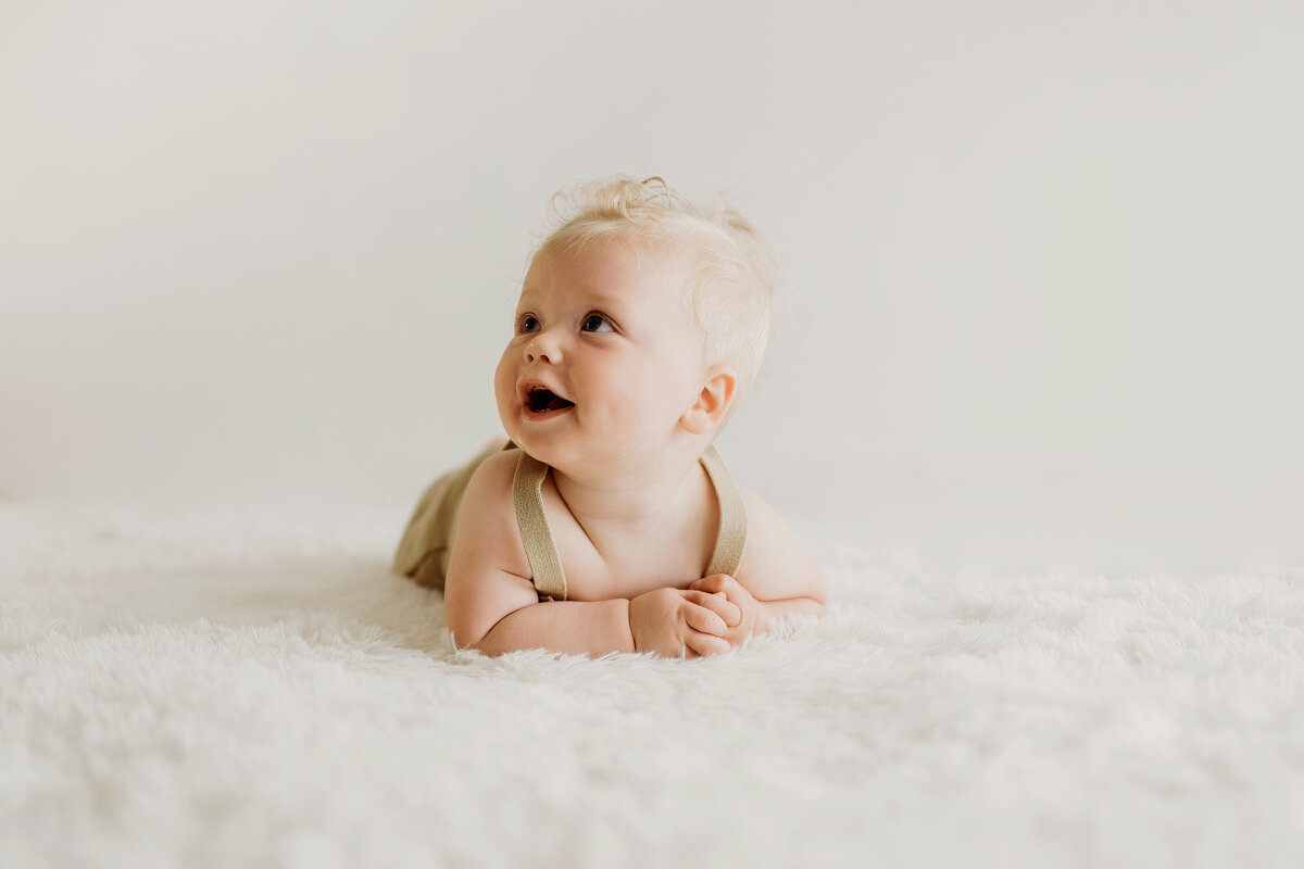 a happy six month old baby boy lays on his stomach in knit overalls on a white rug for his milestone session at an in home Denver studio