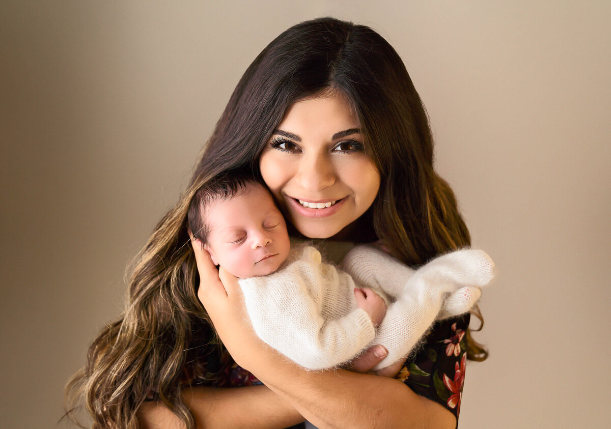 New mom holds her sleeping baby boy and is smiling at the camera.  Mom's cheek is resting against baby's temple. Captured by Bryn Mwar Philadelphia and Main Line photographer Liza Nicole.