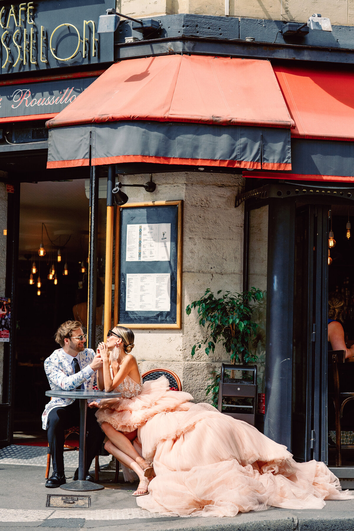 Couple at a cafe enjoying champagne, she in a peach gown with long train at their Paris engagement session, photographed by Italy wedding photographer.