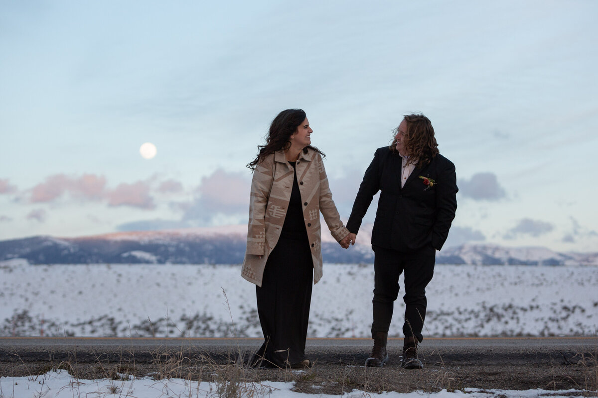 A bride and groom stand on a road in Grand Teton National Park with snowy mountains behind them and the moon peeking out behind some clouds.