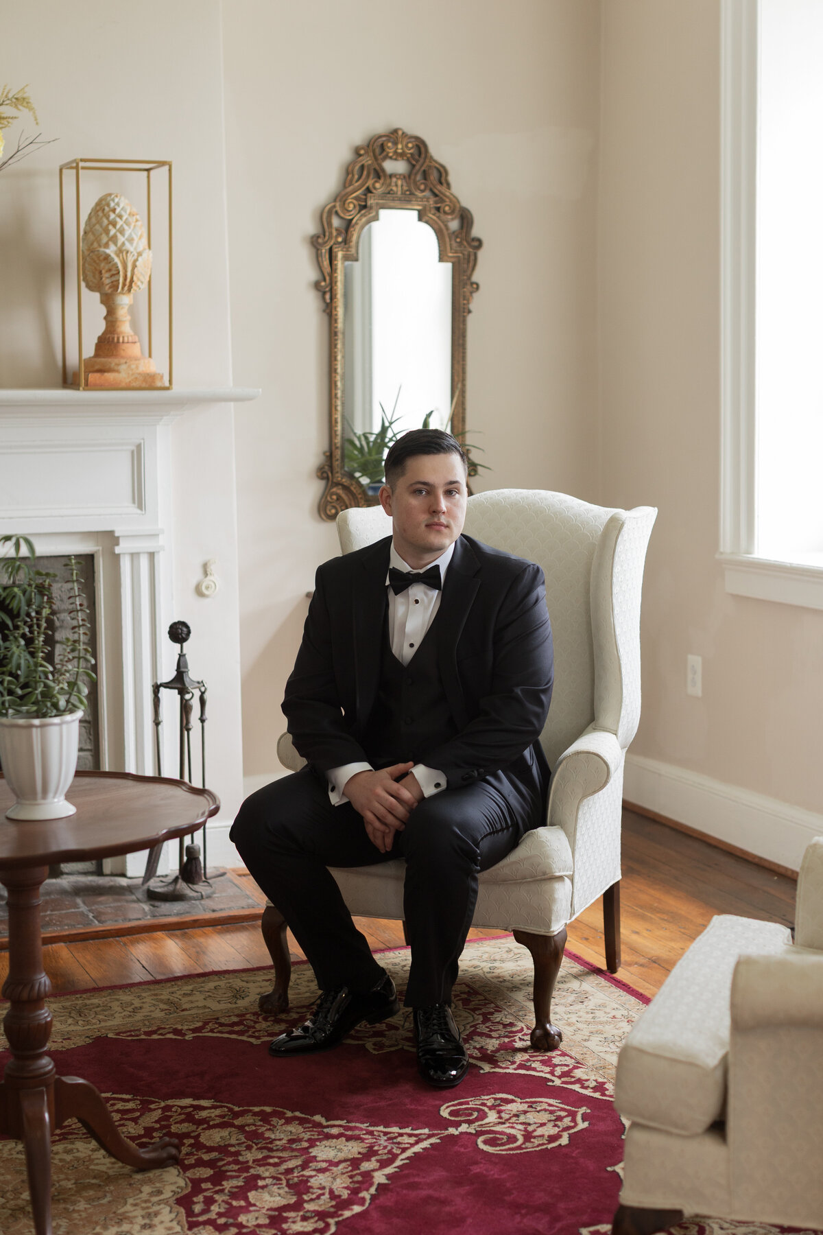 Groom in black tuxedo sitting in vintage armchair at Springfield Manor before spring wedding ceremony.