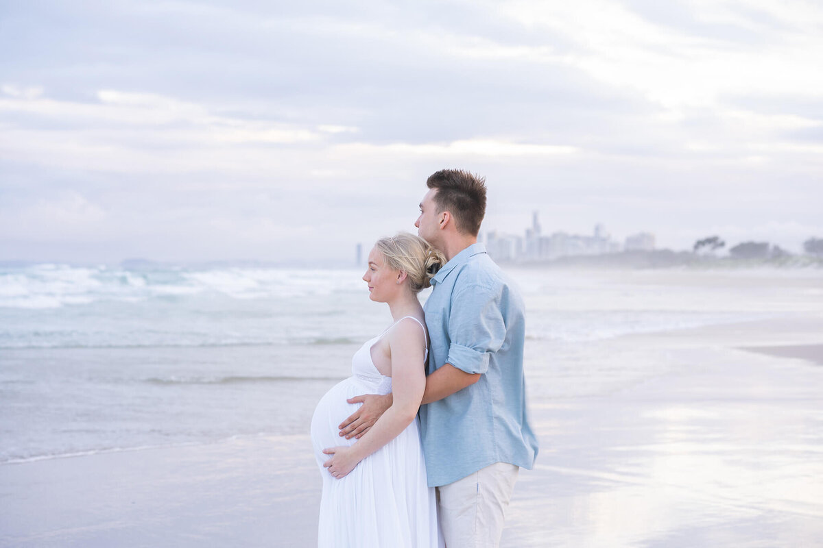 couple looking out into the ocean holding belly bump in golden hour light