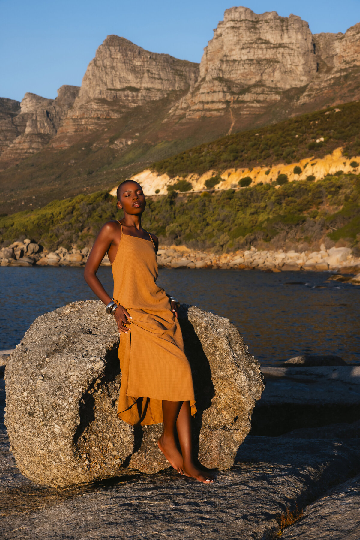 Elegant model leaning barefoot against a large rock by the sea during golden hour, wearing a flowing mustard-yellow dress with bold silver bangles, set against a backdrop of dramatic cliffs and lush greenery in Cape Town.