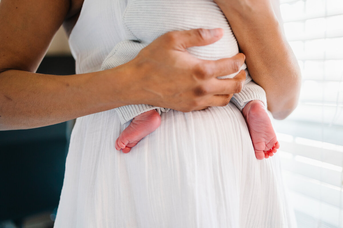 Photo of the feet of a newborn baby being held by his mother in a white dress.