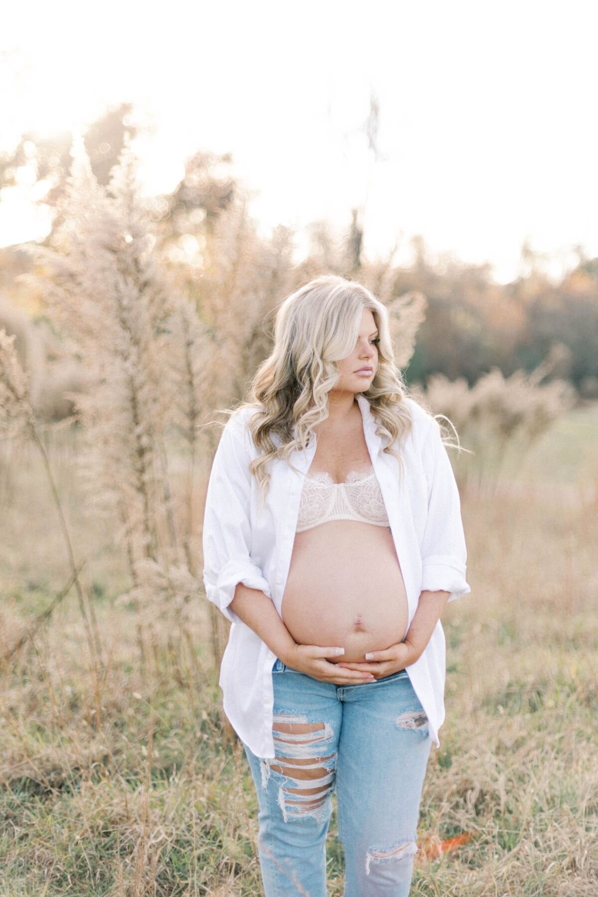 Woman stands in field of tall brush wearing  jeans and white button up opened to show her bare pregnant belly.