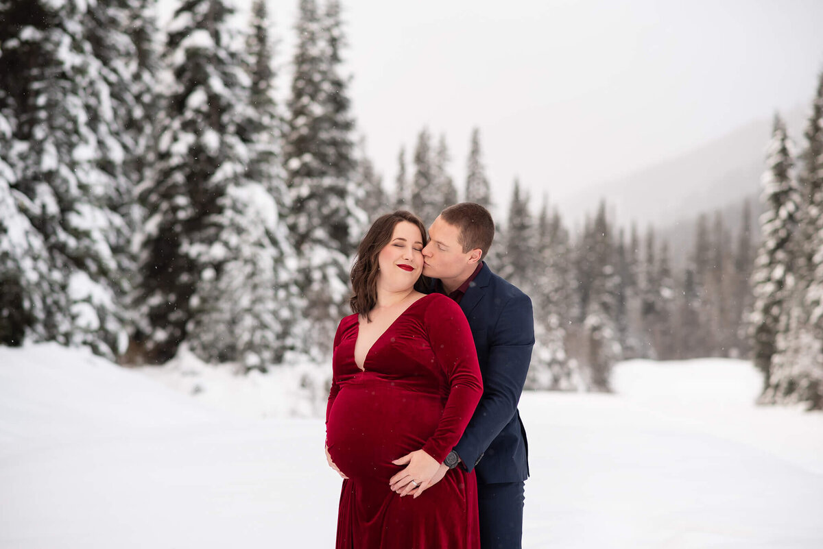 A husband embraces his pregnant wife from behind. She leans into him while he holds her belly. There is a beautiful winter backdrop.  Portrait taken by Brittany Danielle