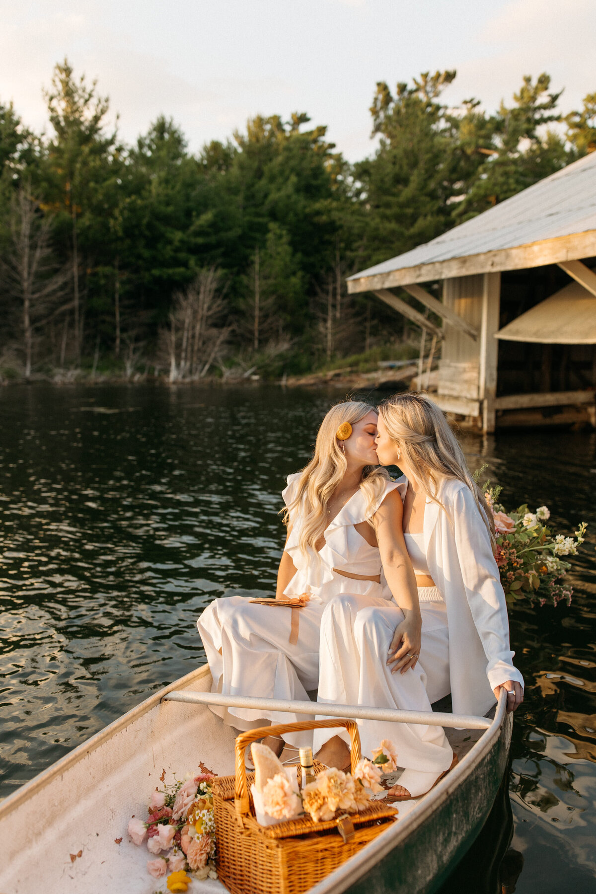 Two women in white dresses sharing a tender moment on a canoe adorned with flowers on a serene lake