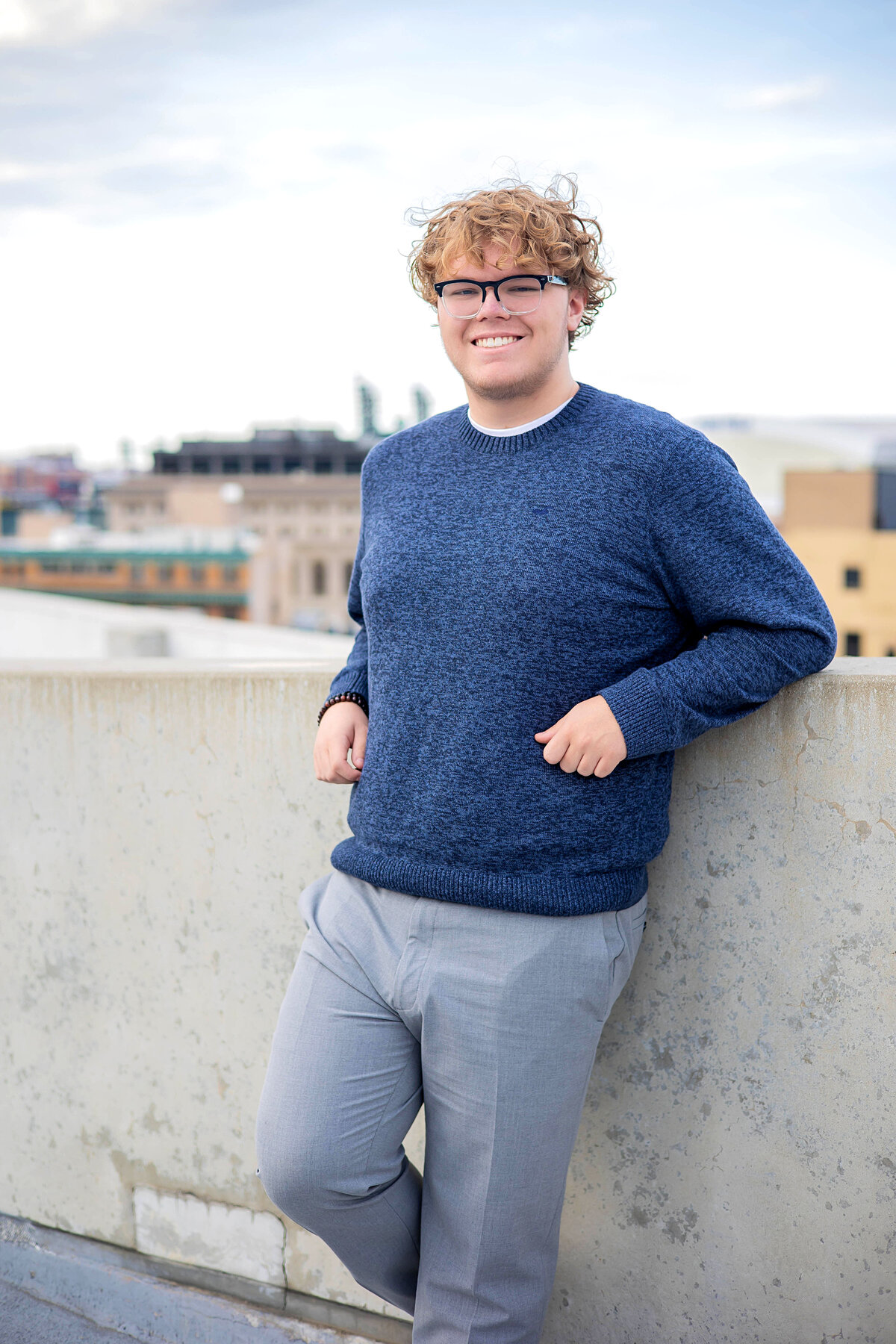 Caucasian senior boy wearing a blue sweater, gray dress, and brown casual shoes standing against a brick wall posing casually