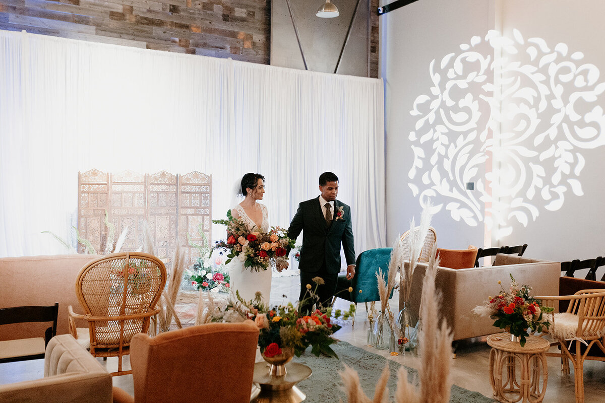 The bride and groom walk through a lounge area decorated with vintage chairs, pampas grass, and vibrant floral arrangements, with an illuminated pattern on the wall behind them.