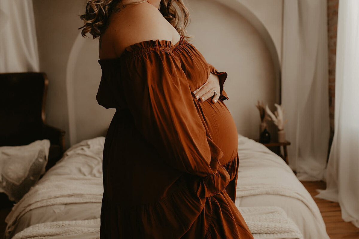a pregnant woman holds her bump wearing a brown off the shoulder dress in a studio in rochester, ny