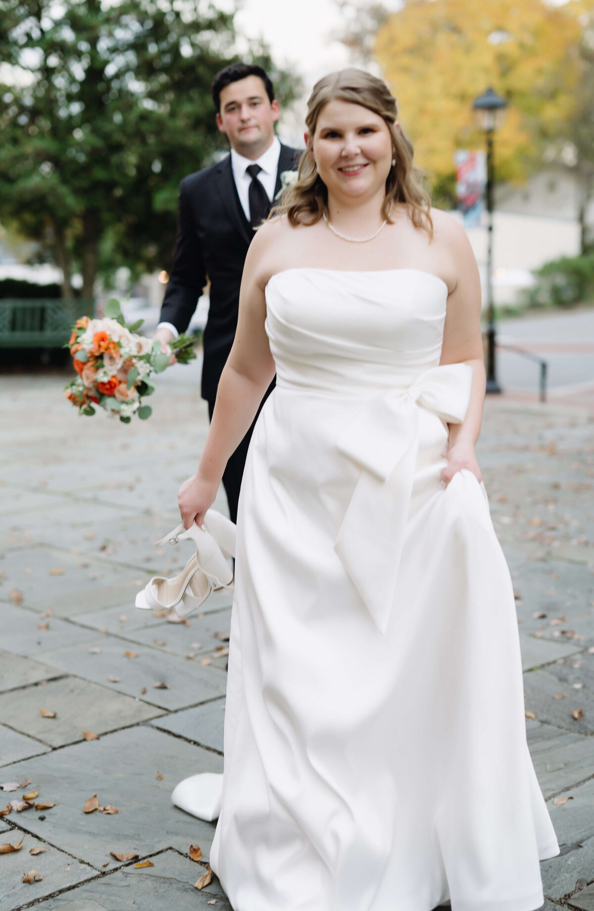 bride holding her wedding shoes and holding her wedding dress with her other hands as she walks across a stone patio smiling with her groom trailing behind her at middleburg community center