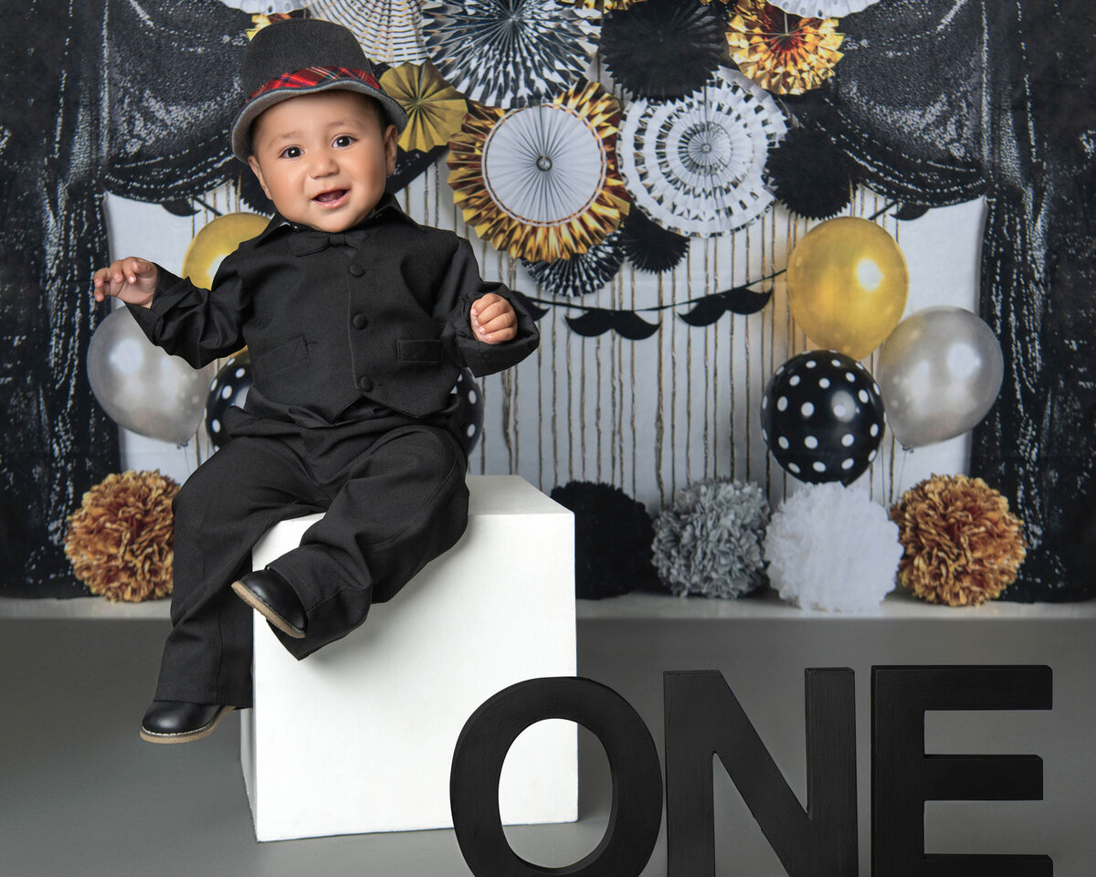 A happy baby sits on a white box for its birthday in black pants, shirt and hat in an Orange County milestone photography studio