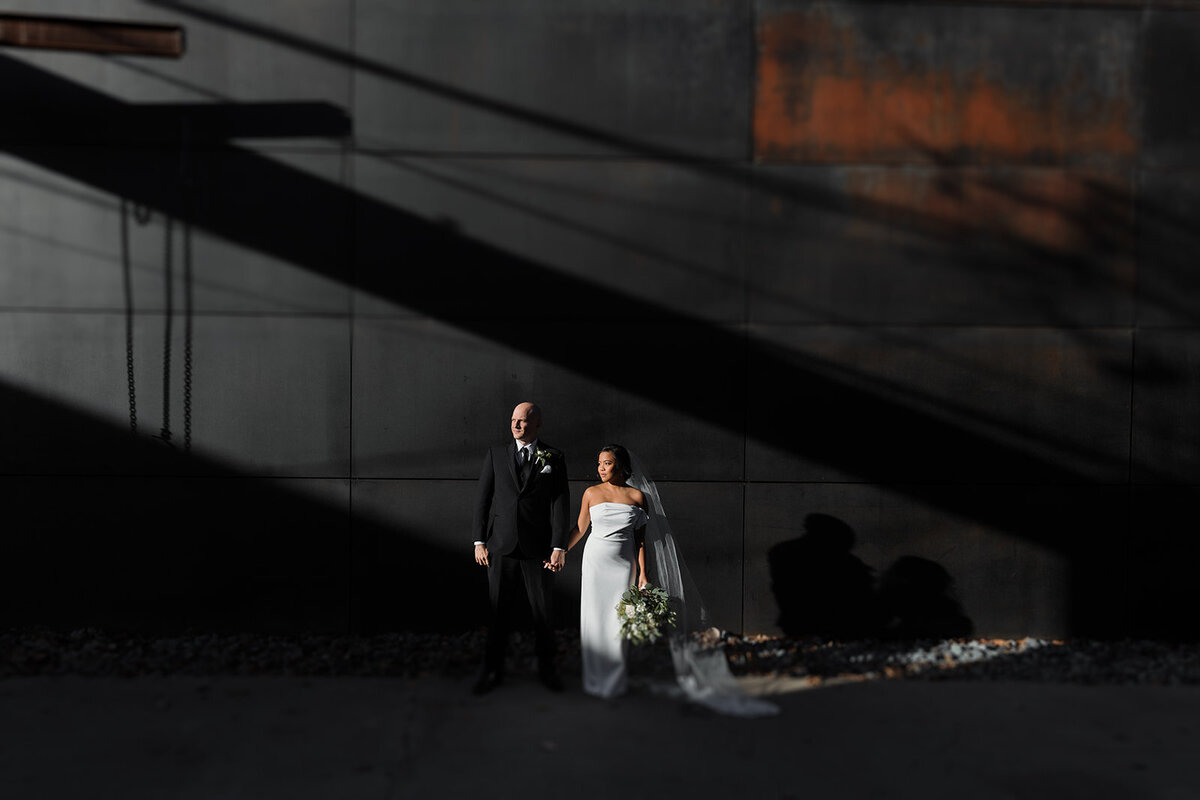 couple stands in bright sun with shadows on dark wall behind them. They hold hands and look into sun.