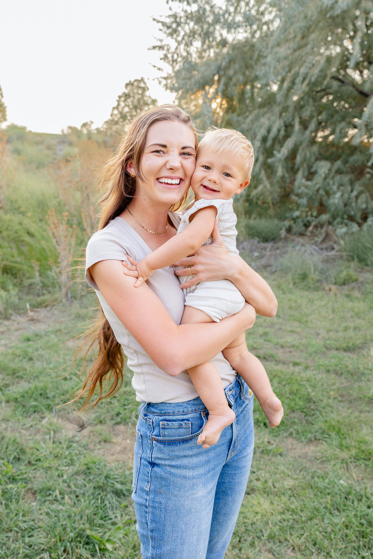 A joyful mother holds her baby boy close, both smiling brightly in a picturesque outdoor location in Eagle Mountain, Utah.
