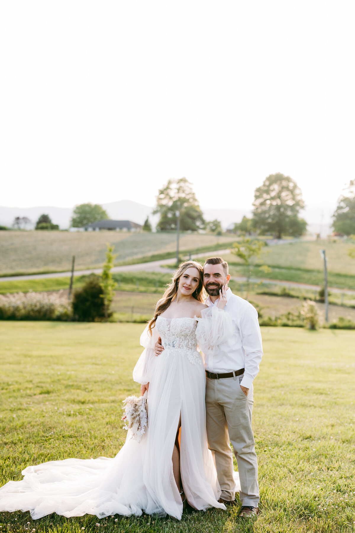 wedding portrait by Virginia wedding photographer with bride and grom on a Charlottesville wedding venues lawn posing with the bride holding her grooms jawline as she leans her back into him and he holds her waist with rolling hills behind them at sunset