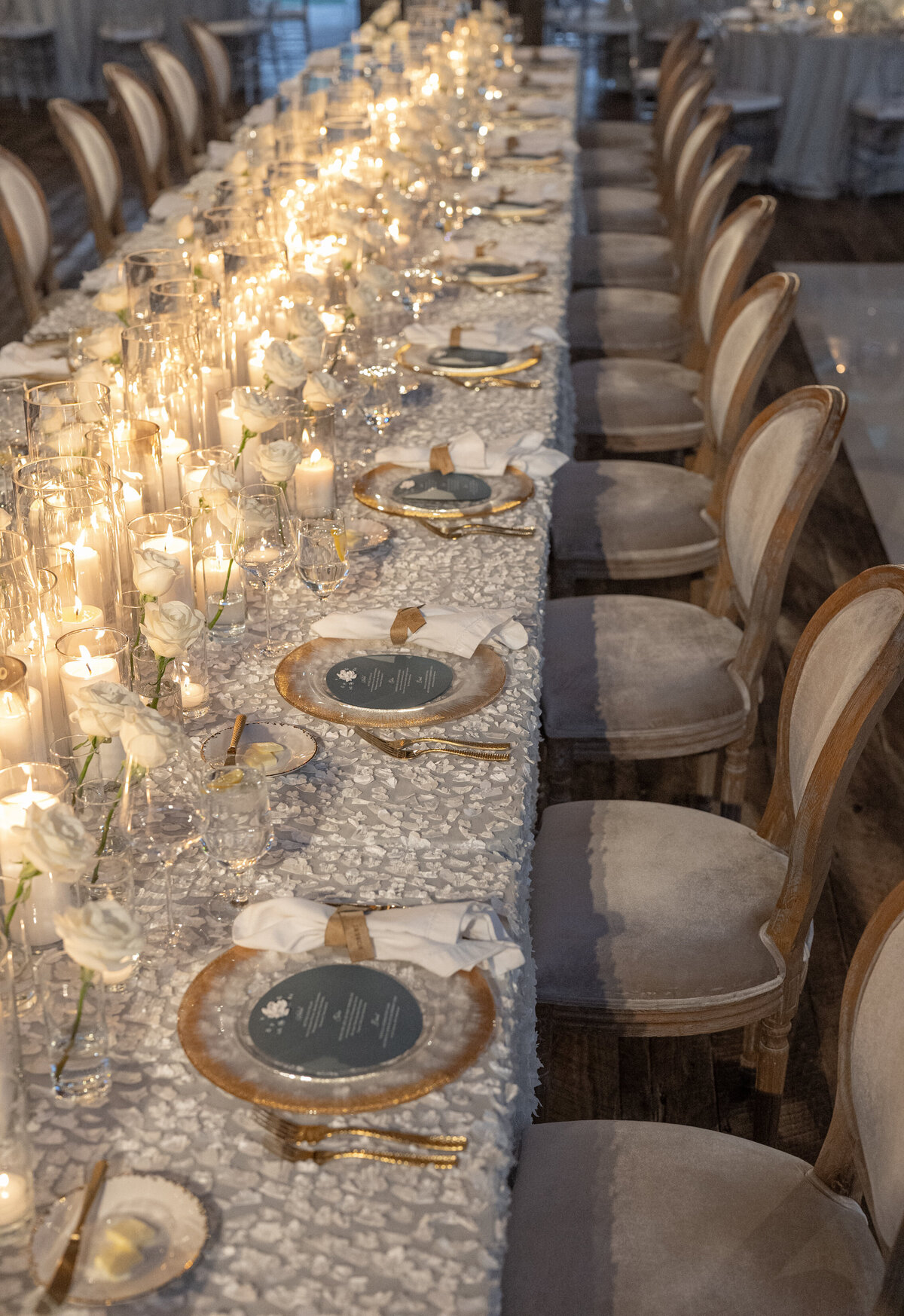 Textured floral linens, with gold lined flatware and dozens o white candles and white roses