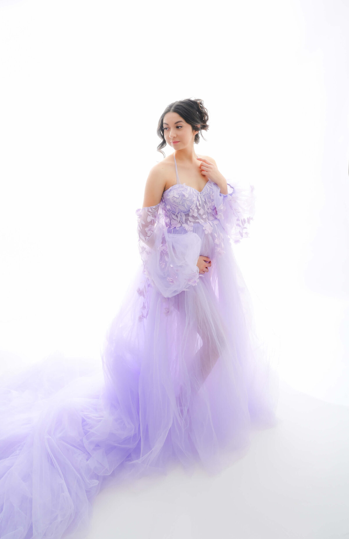 Beth Benecki of E. Benecki Photography photographs a backlit maternity photo session with a white background.  Expecting mother is wearing an off the shoulder chiffon lavender dress with a long train.