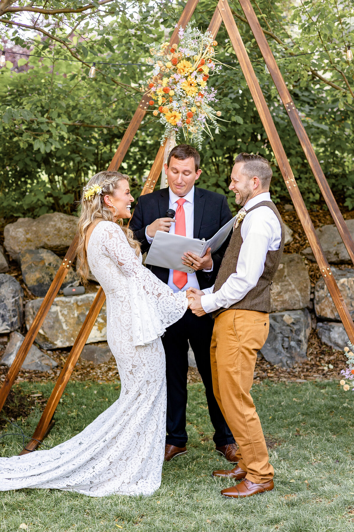 The bride and groom hold hands and laugh together during their wedding vows in front of a triangle wooden arch adorned with yellow and orange flowers, set against the trees and rocks of Provo Canyon.