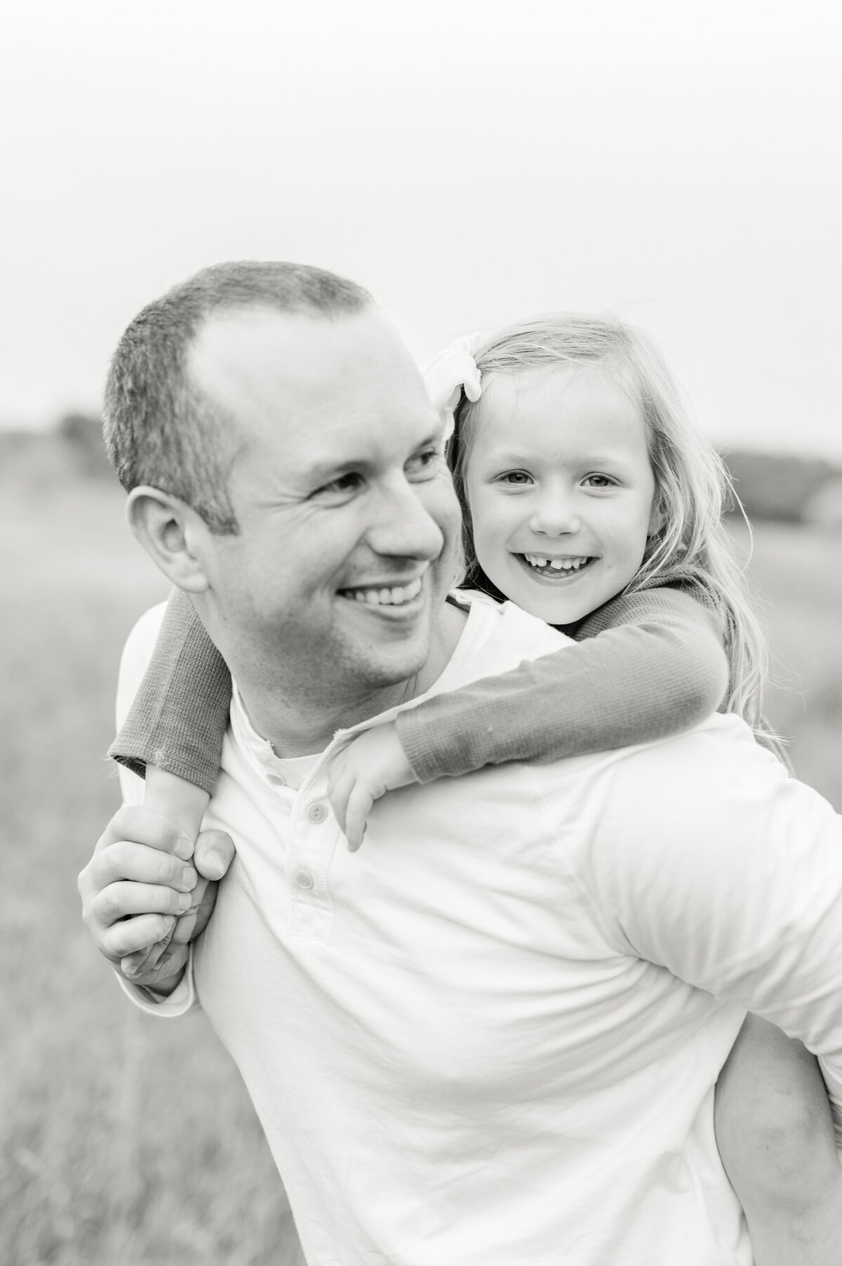 Black and white image of a dad giving his daughter a piggy back ride while smiling back toward her