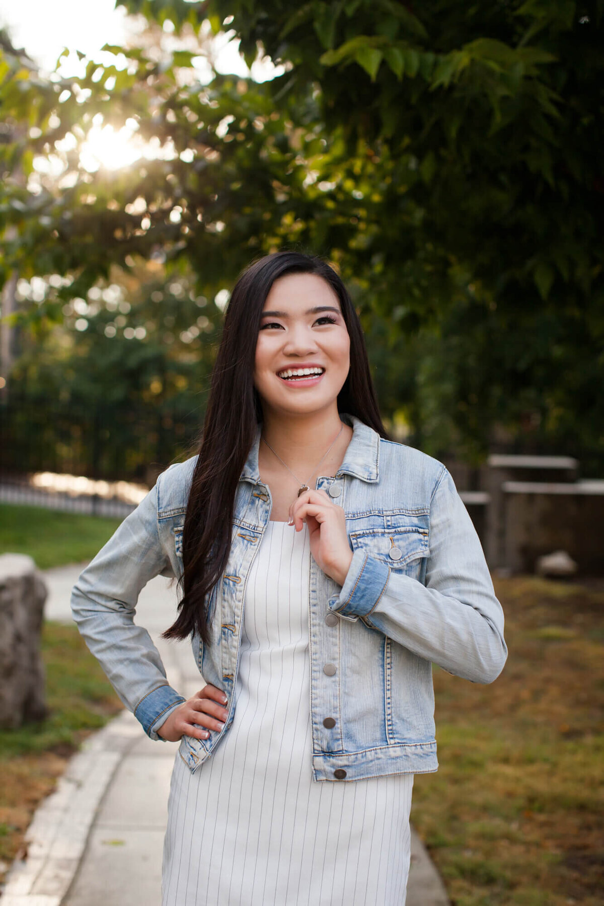 A laughing Asian American teen poses for a senior picture wearing a striped short white dress and jean jacket. Captured by Springfield, MO senior photographer Dynae Levingston.