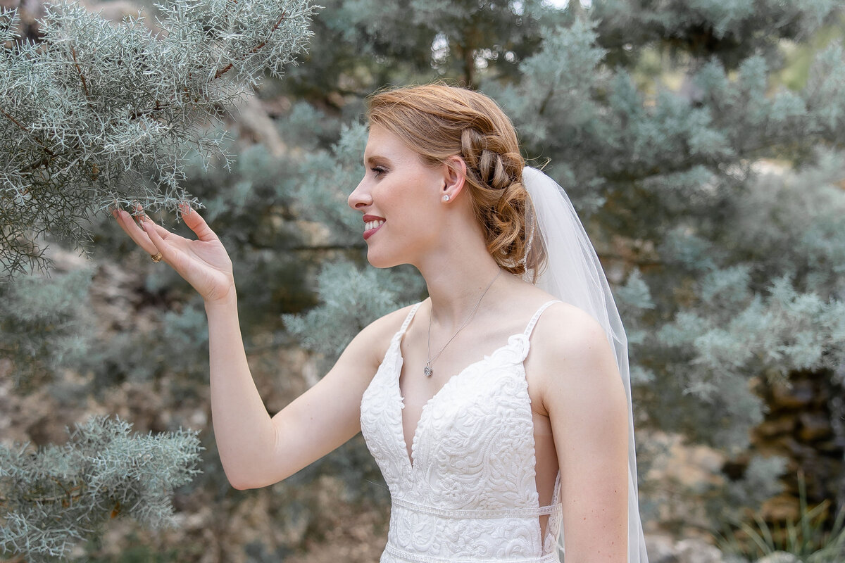 bride touches pine tree branch and smiles in spaghetti strap wedding dress and veil