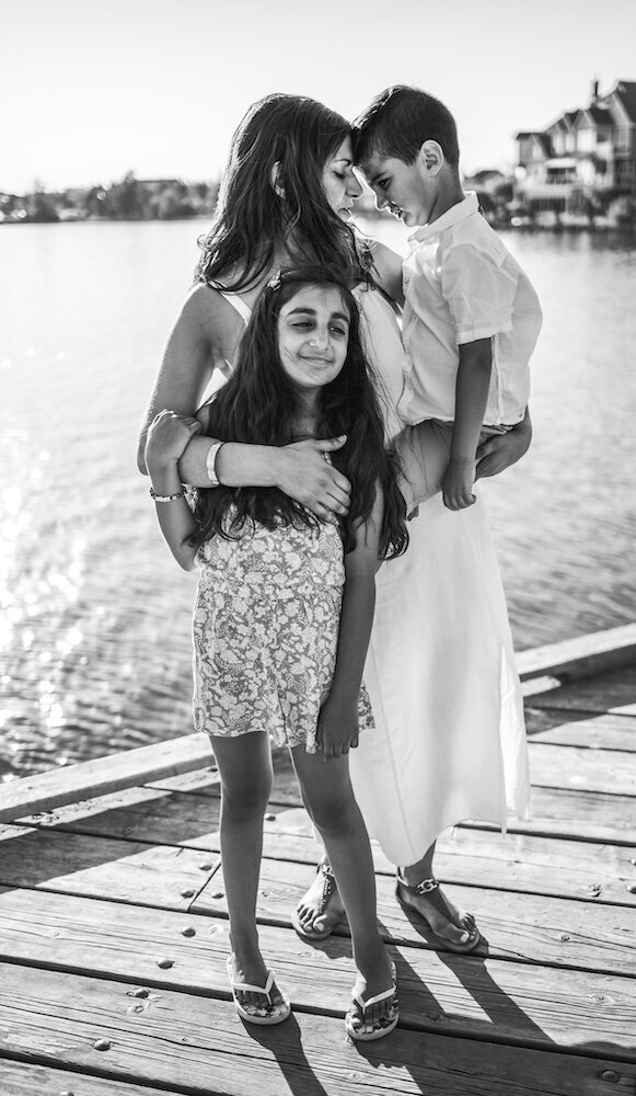 A mom in a white dress is holding her son on her hip and hugging her daughter with her other hand