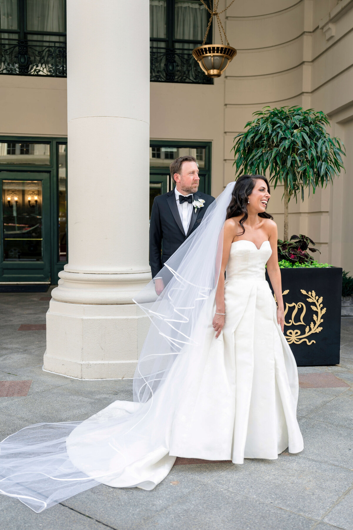 A bride in a white wedding gown and long veil smiles and looks to her right, standing beside a groom in a black suit and bow tie. They are outside near large pillars and a potted plant. The setting appears elegant and formal.