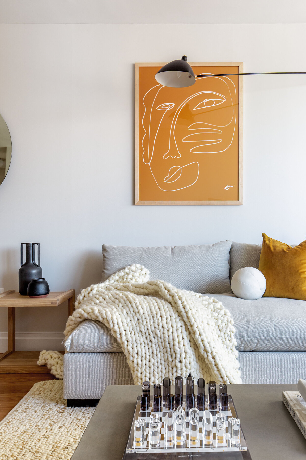Living room with grey armless sofa, yellow abstract artwork on wall, serge mouille style double sconce light, leather armchair, wooden side table, and concrete coffee table with books and decor stacked on top