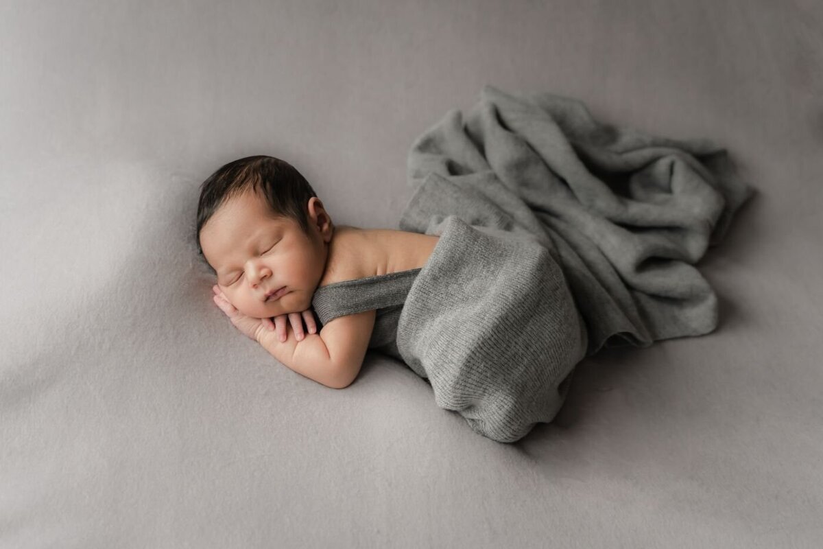 Newborn baby in a gray wrap, sleeping peacefully on a light gray backdrop with a matching blanket.