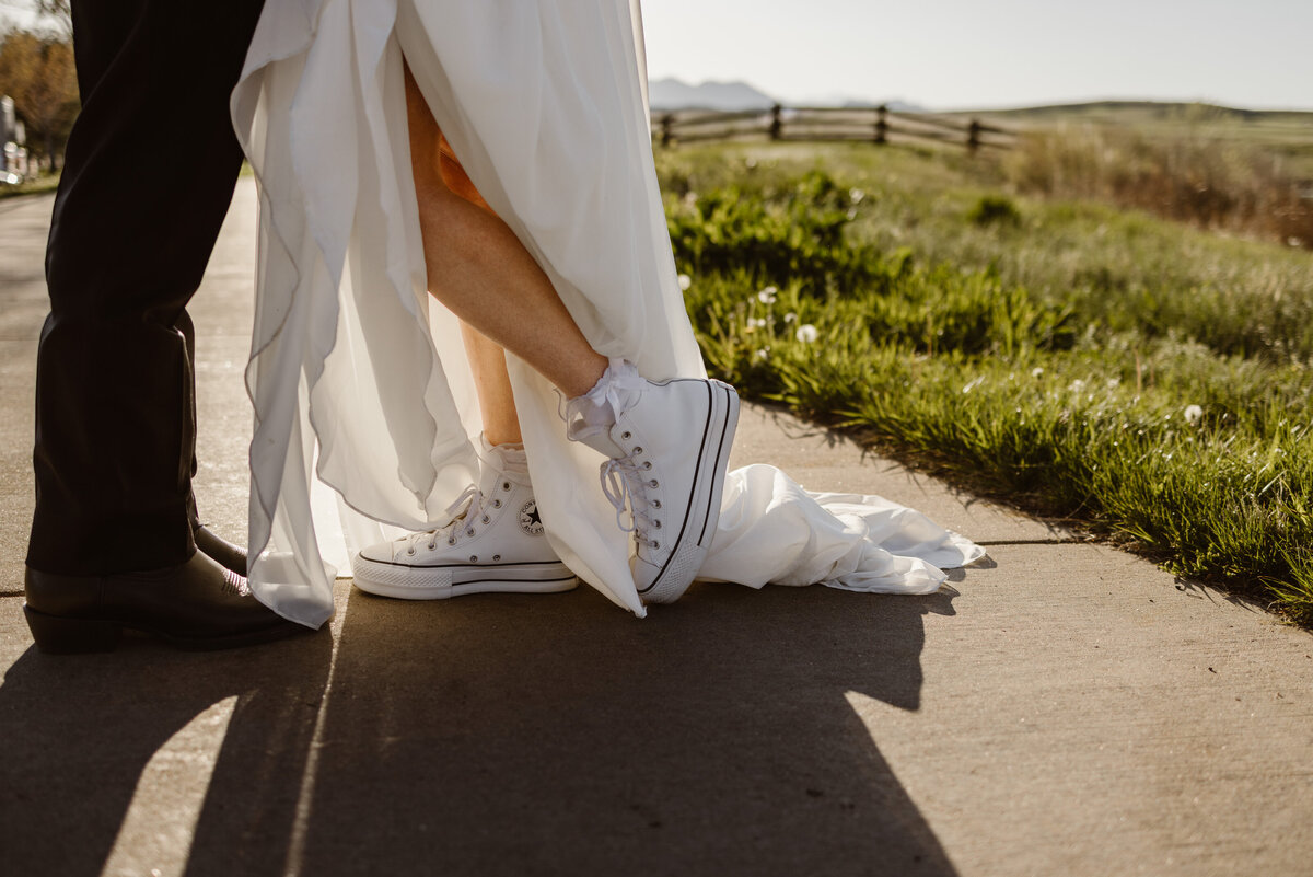 Close-up of a bride wearing white Converse sneakers and frilly socks under her flowing wedding dress, gently leaning into her partner on a sunlit path.