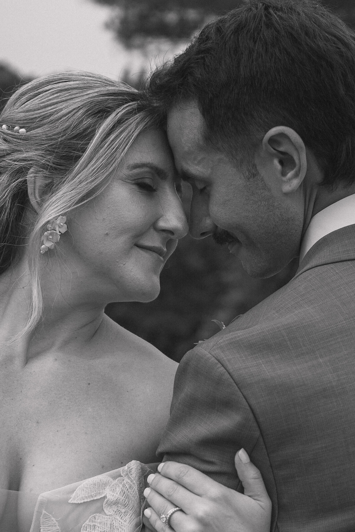 Close-up of a bride and groom touching foreheads and smiling with eyes closed. The bride, adorned with delicate earrings and a floral lace gown, shows her wedding ring as she gently holds the groom's arm. The groom wears a light suit and has a mustache.