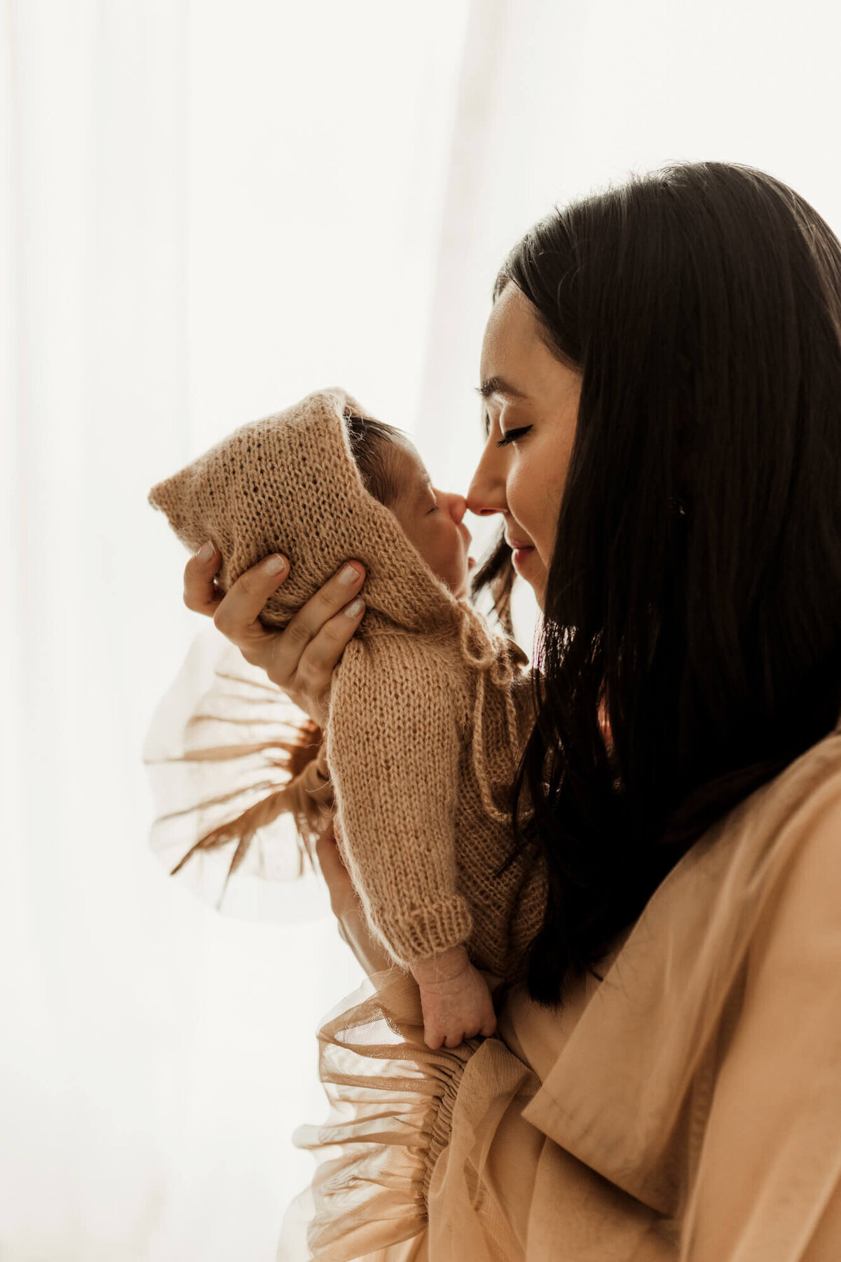Mother touches her nose to her newborn baby boy's nose with a window behind them.