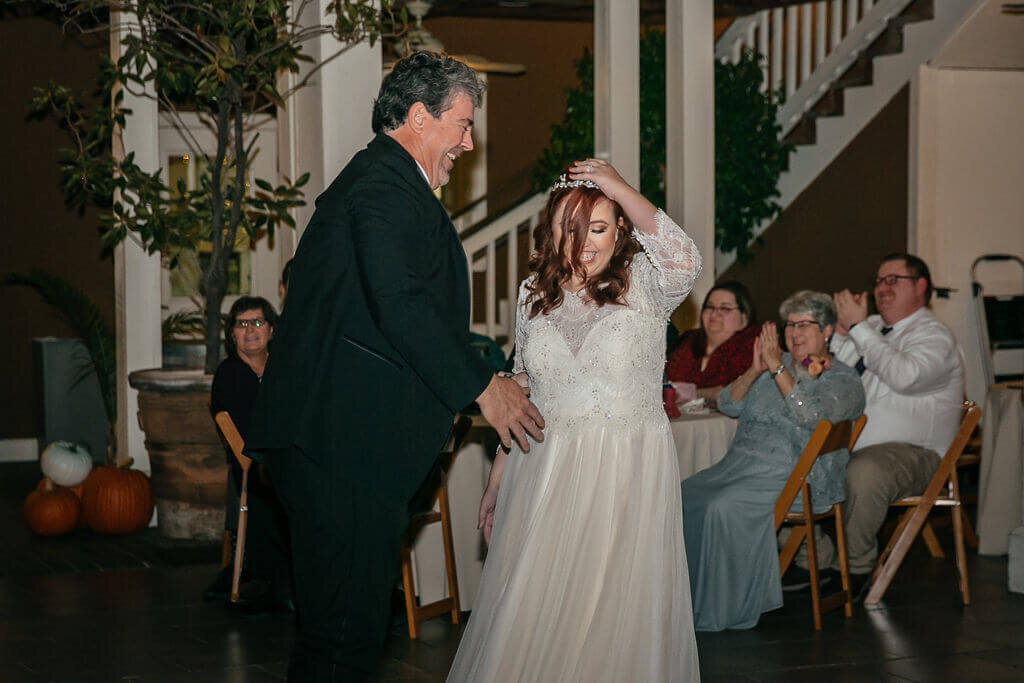 A bride in a white gown dances with a man in a suit, both smiling. Seated guests watch and clap, with a staircase and pumpkins in the background. 