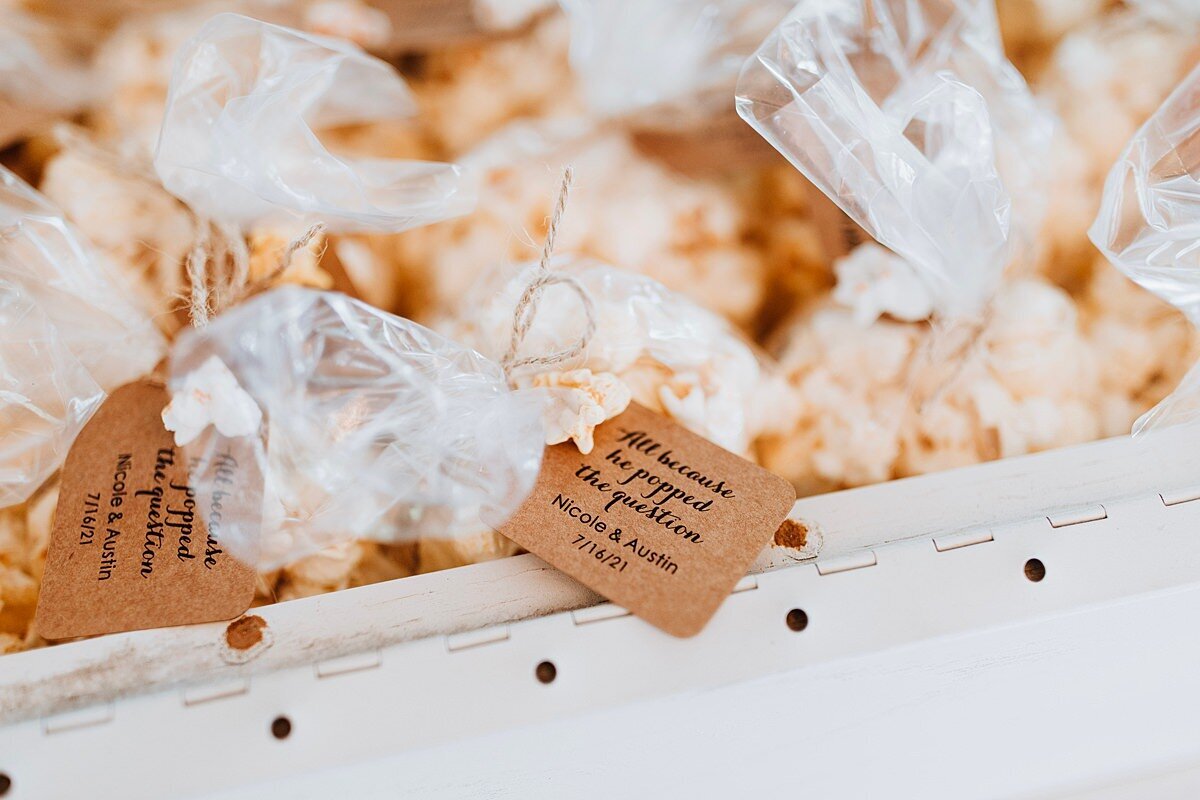 A white farm table filled with mini bags of popcorn with small brown tags. Popcorn wedding favors at Steel Magnolia Barn
